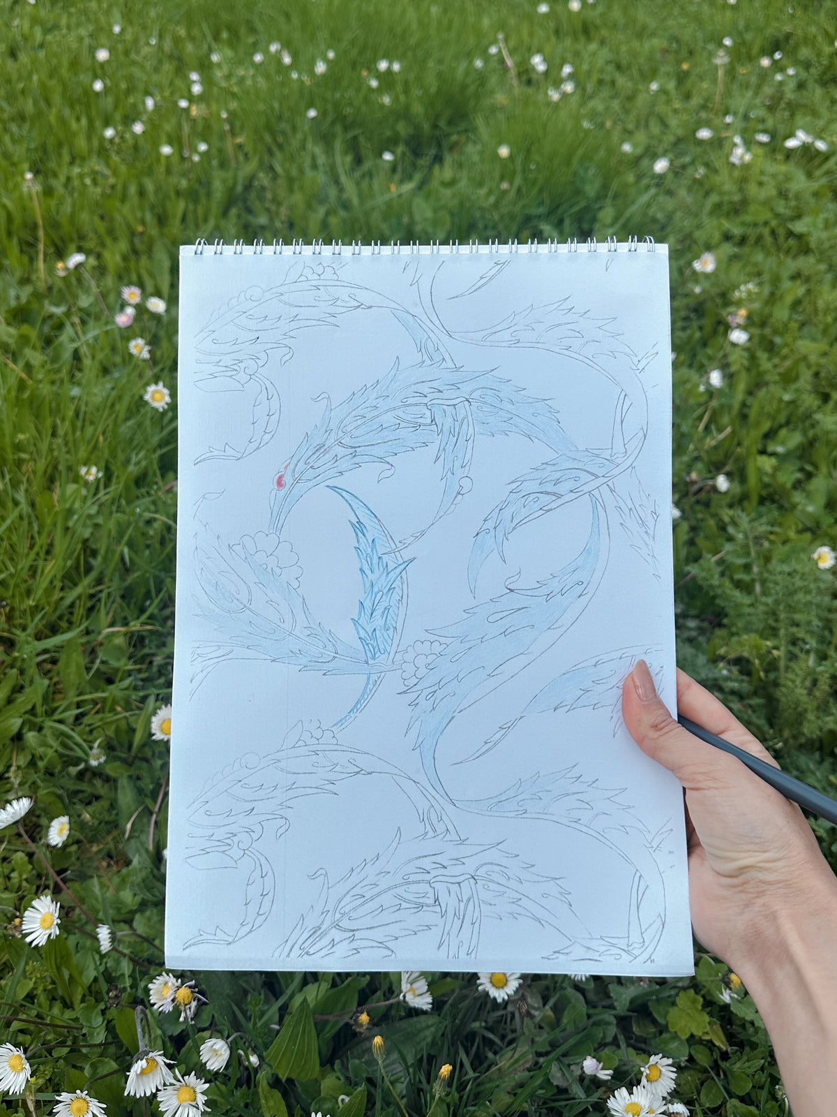 A hand holds a spiral-bound sketchbook showing a detailed blue line drawing of a Turkish traditional leaves pattern, set against a real outdoor grassy field with small white flowers.