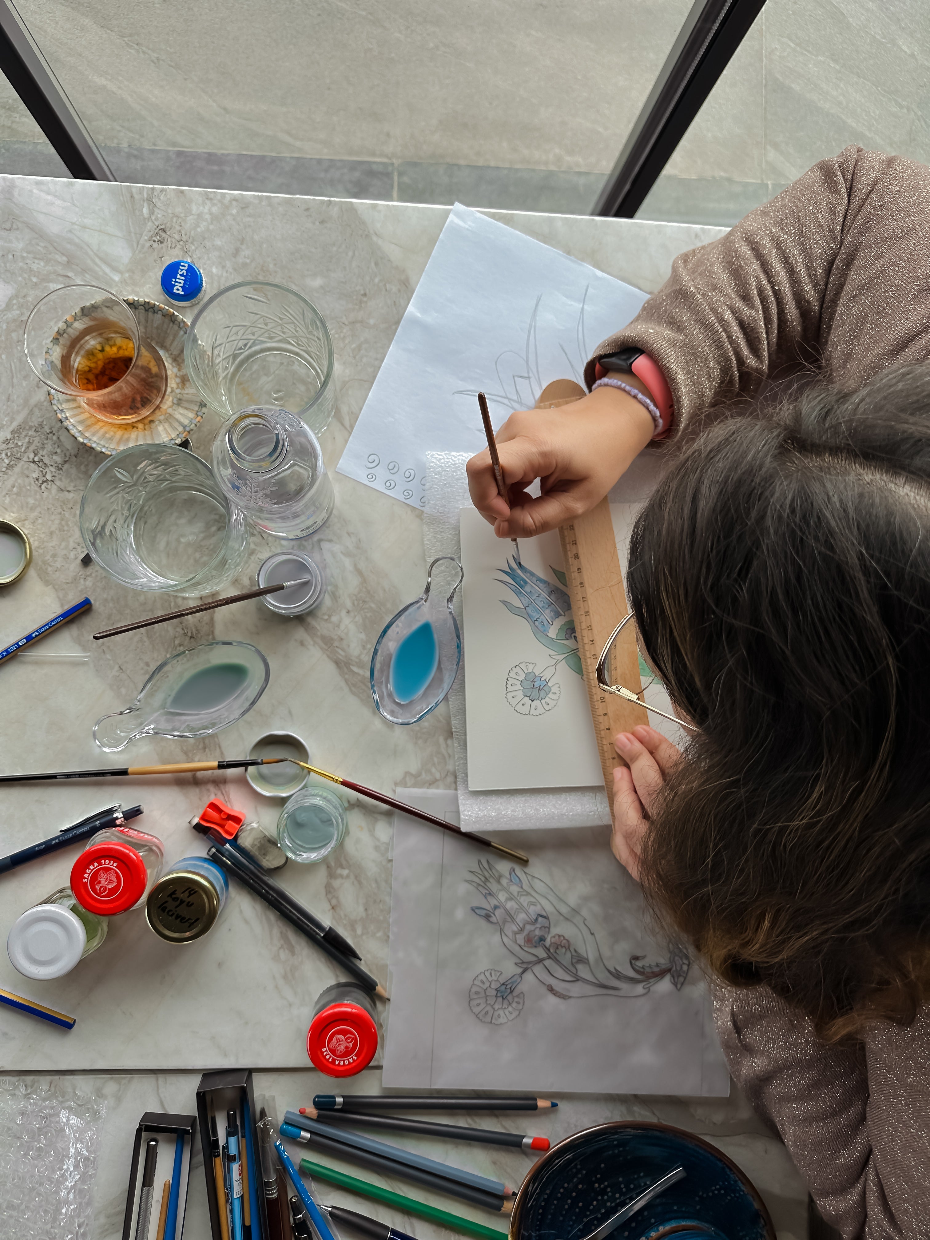 A woman sitting on the table and is coloring the ceramic tile with blue color, holding a small sized brush.